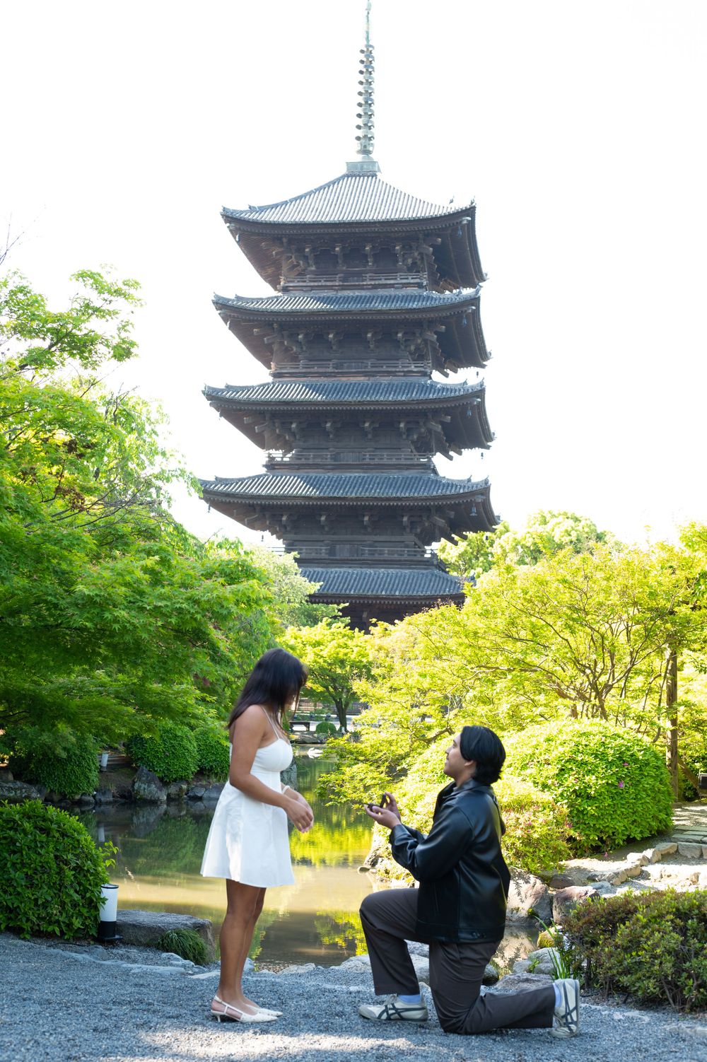 Man kneeling in a marriage proposal to a woman in a white dress beside a pond, with a historic five-story pagoda and green trees behind.