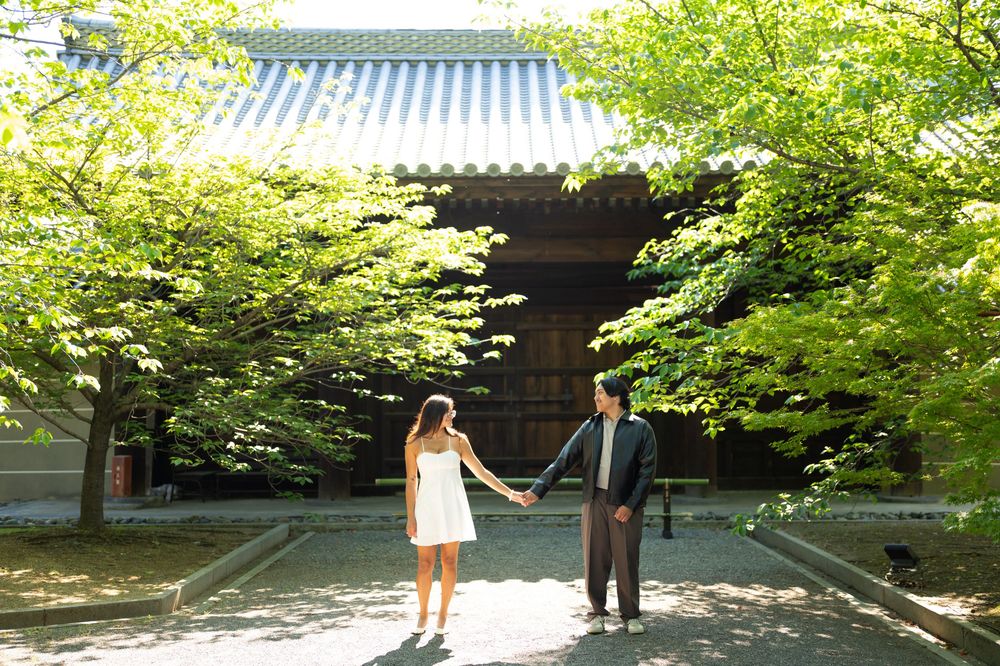 Couple holding hands on a stone path in a sunlit courtyard with green trees and a traditional Japanese-style building behind them.