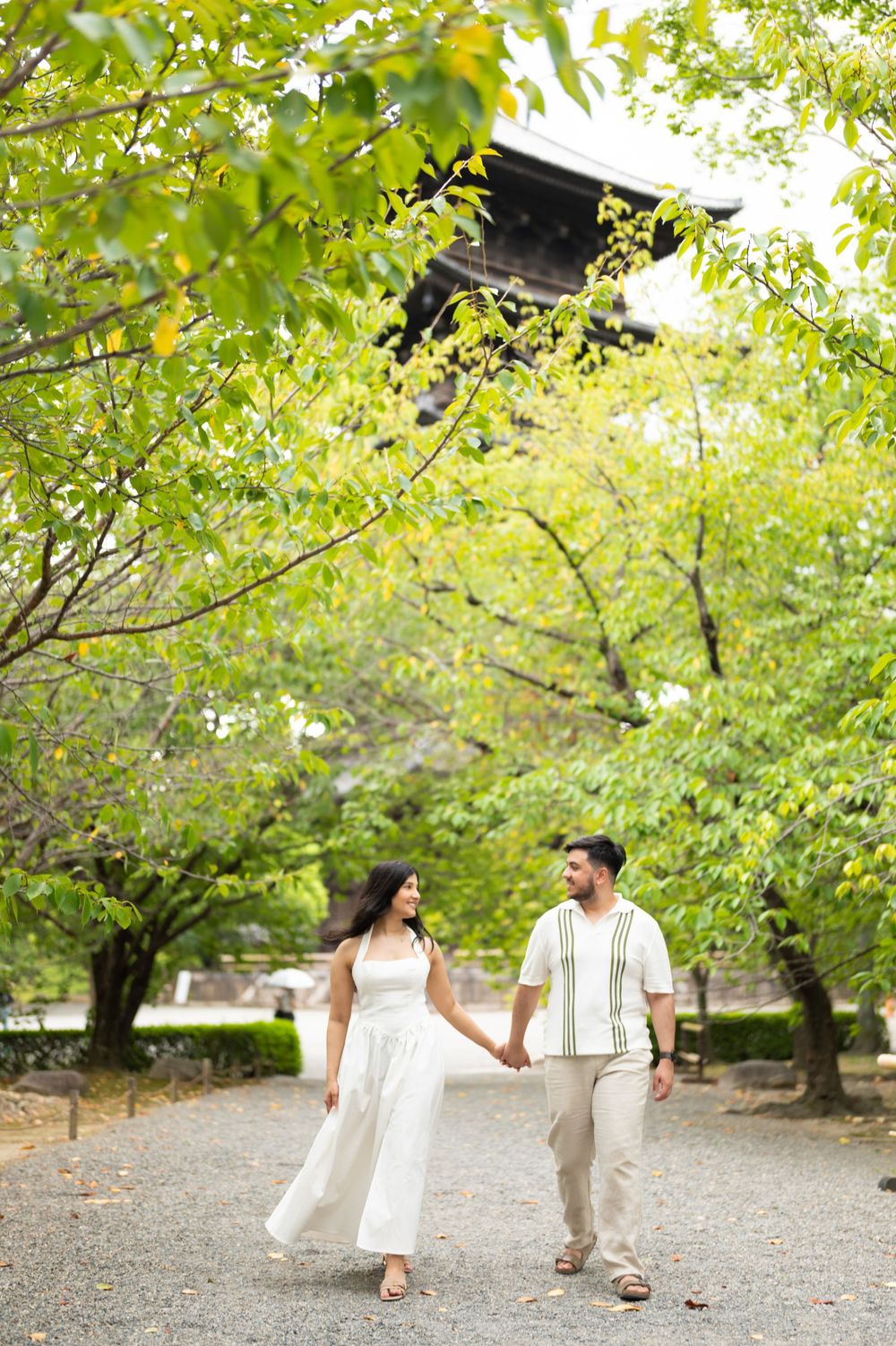 Couple in white and beige outfits holding hands while walking along a gravel path beneath green trees near a traditional Japanese temple.