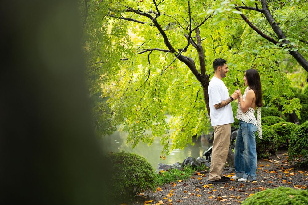 Couple holding hands and facing each other on a garden path beneath lush green trees with sunlight filtering through the leaves.