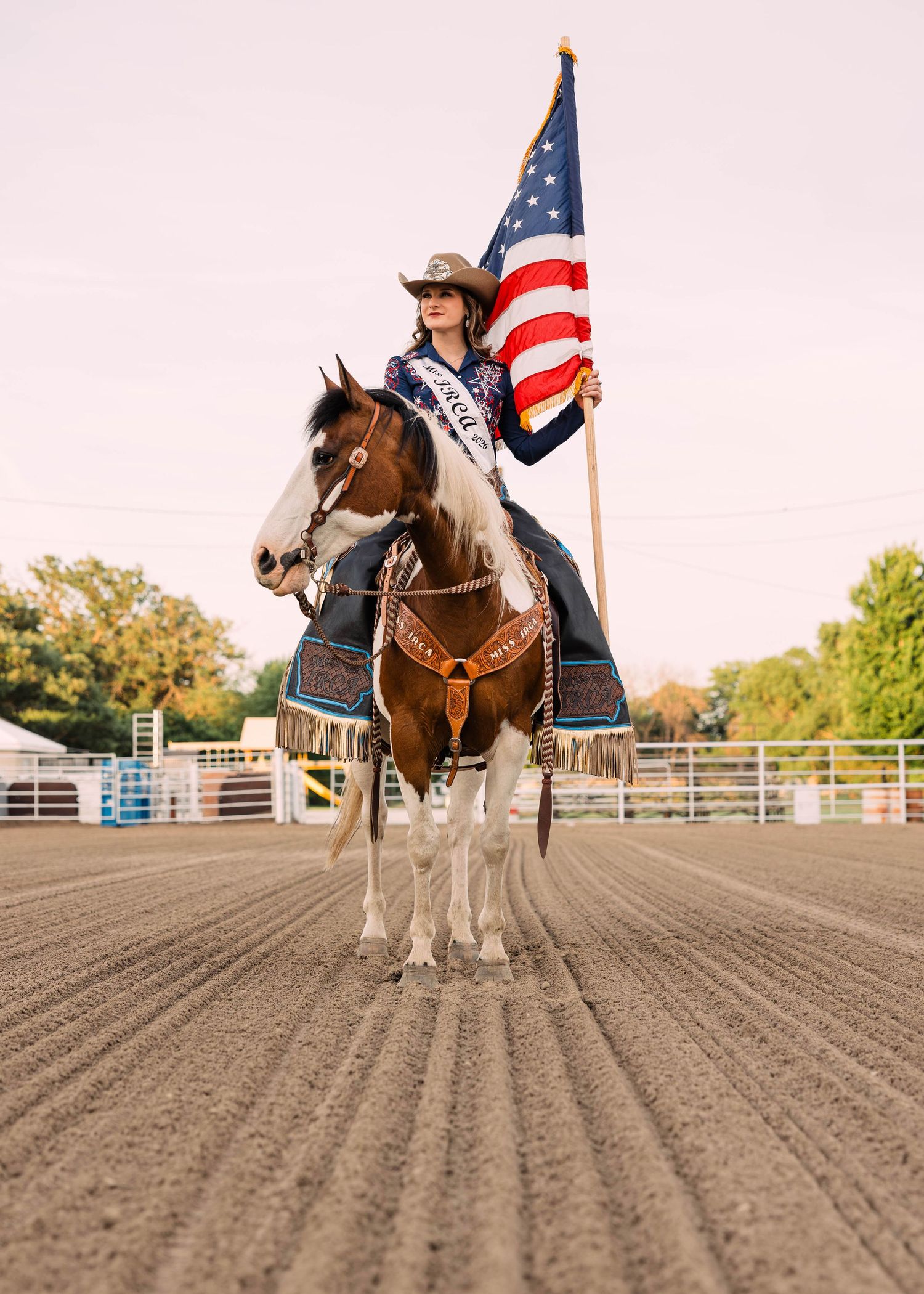 Rodeo queen in a cowboy hat and sash rides a pinto horse in an arena, carrying an American flag at sunset.