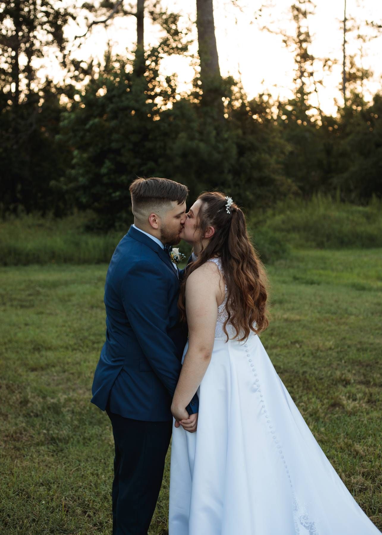 Smiling groom in a navy suit and boutonniere embraces his bride beneath her sheer veil in warm golden sunset light.