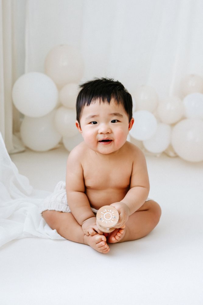Black and white photo of a toddler in a linen outfit sitting on a wood floor playing with a wooden toy cake by white balloons.