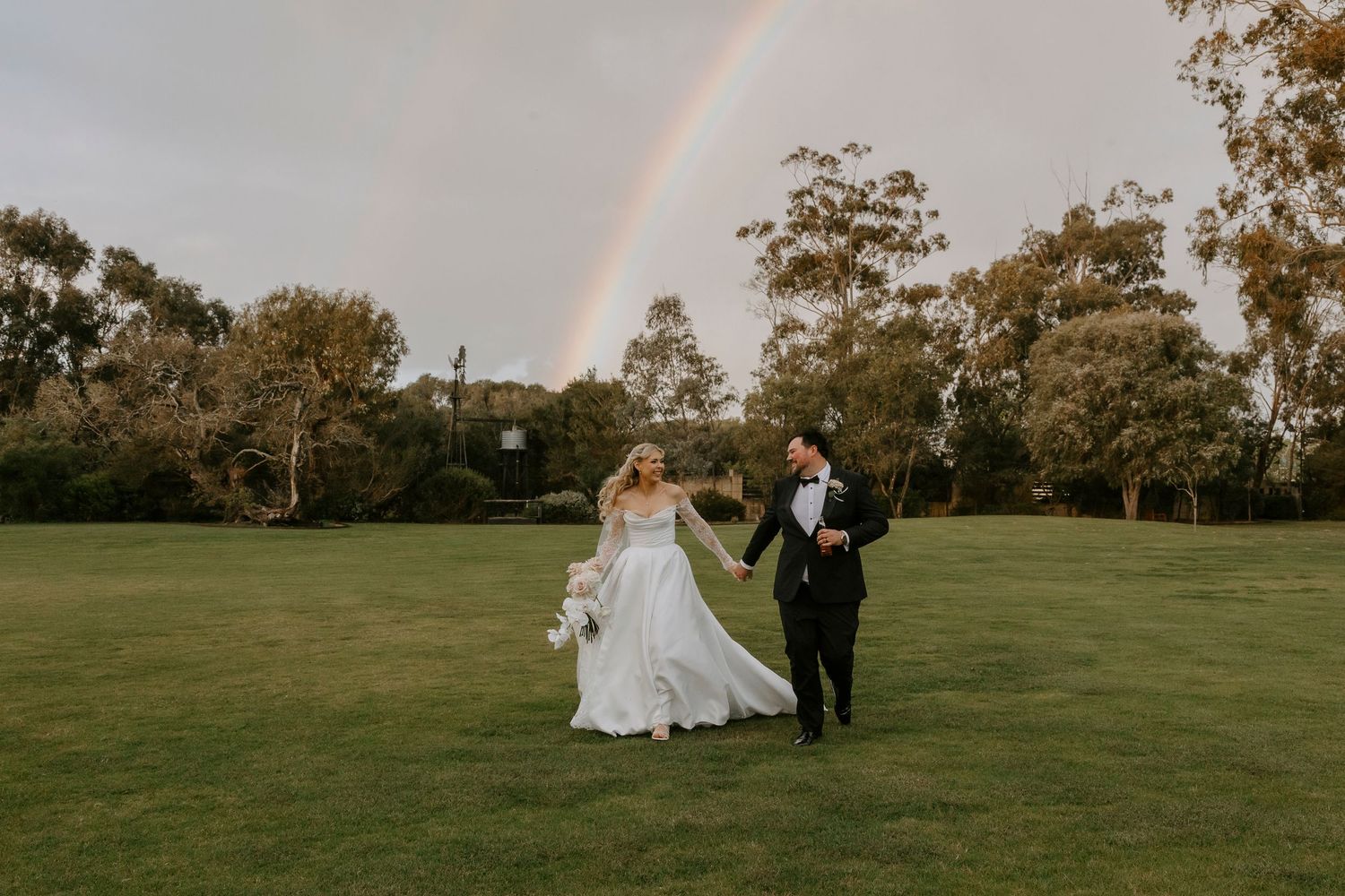Bride in a white gown holding a bouquet walks hand in hand with groom in a black suit across a green lawn beneath a rainbow.
