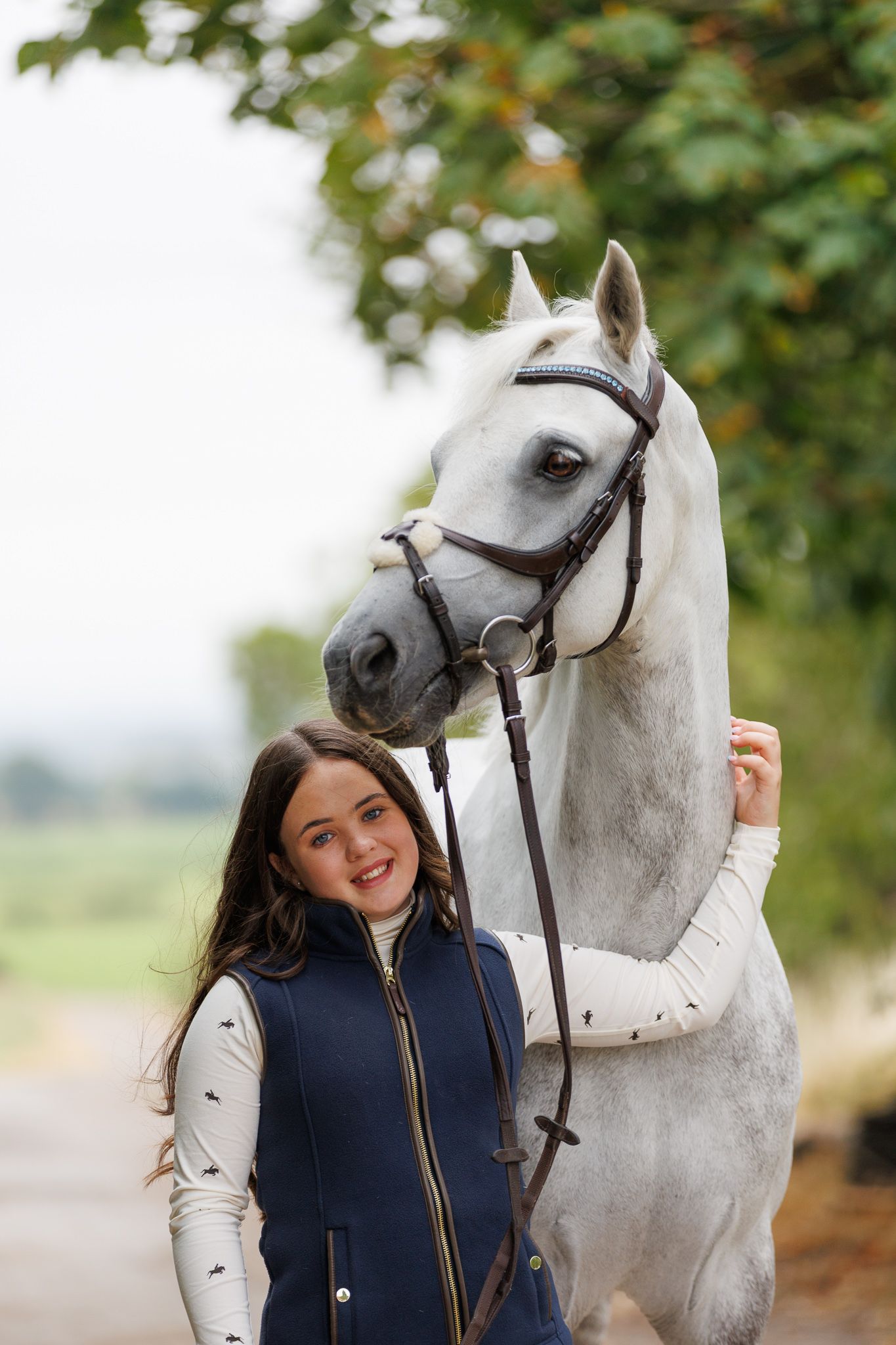 A smiling young woman in a navy vest stands beside a white horse wearing a brown bridle outdoors.