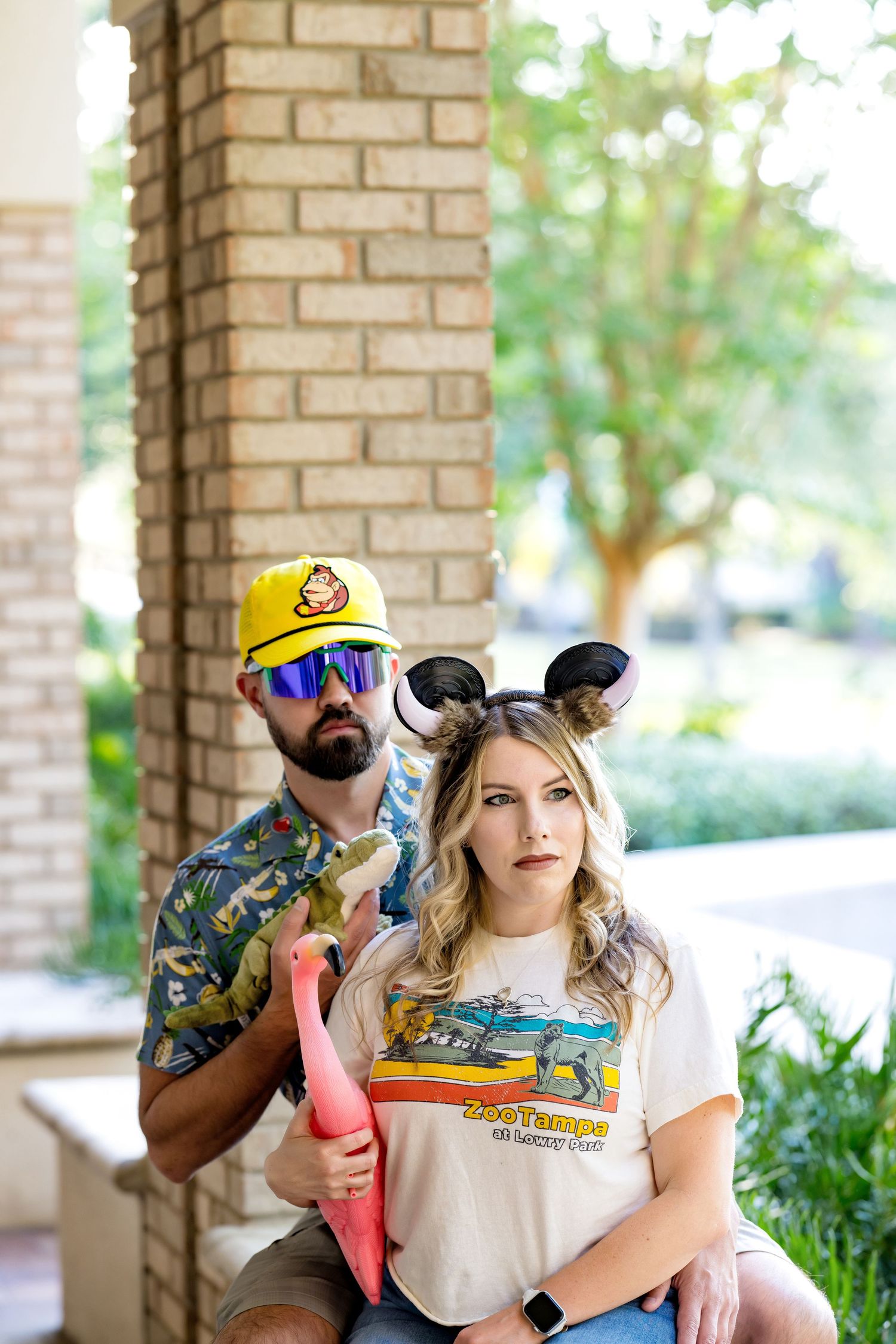 Bearded man in tropical shirt, yellow cap, and mirrored sunglasses holding green vegetable by ornate column outdoors.