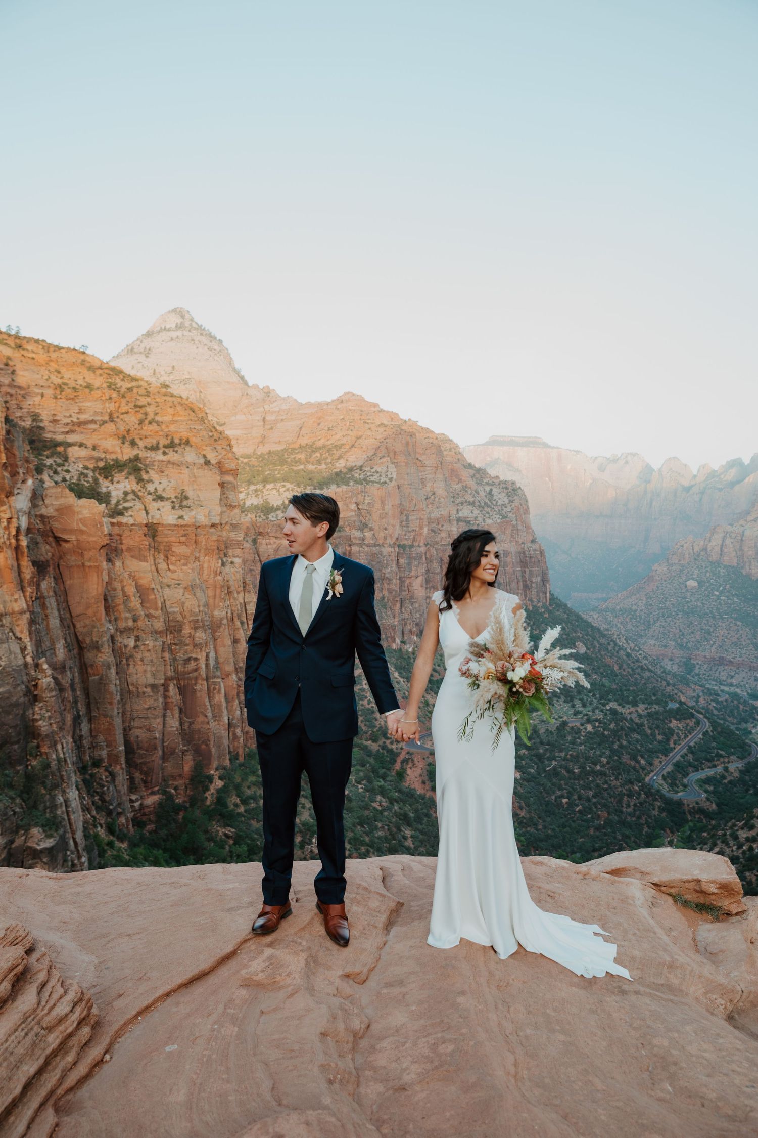 Bride and groom hold hands on a rocky overlook at Zion National Park, with stunning red canyon views behind them.