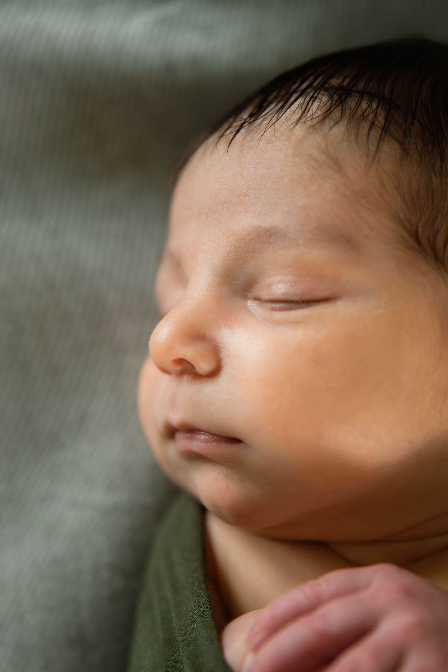 Peaceful newborn baby sleeping soundly, wrapped in green cloth, with dark hair and delicate features in soft light.