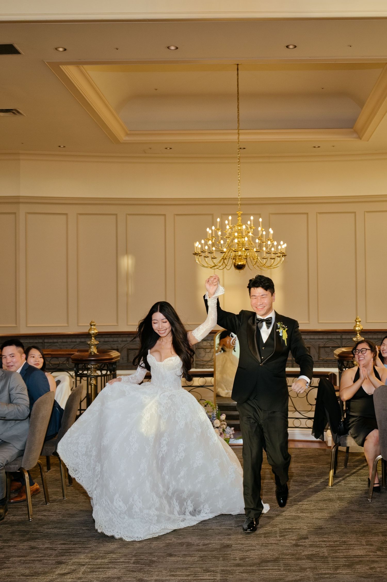 Bride in a lace ball gown and groom in a black tuxedo hold raised hands as they enter a wedding reception under a chandelier.