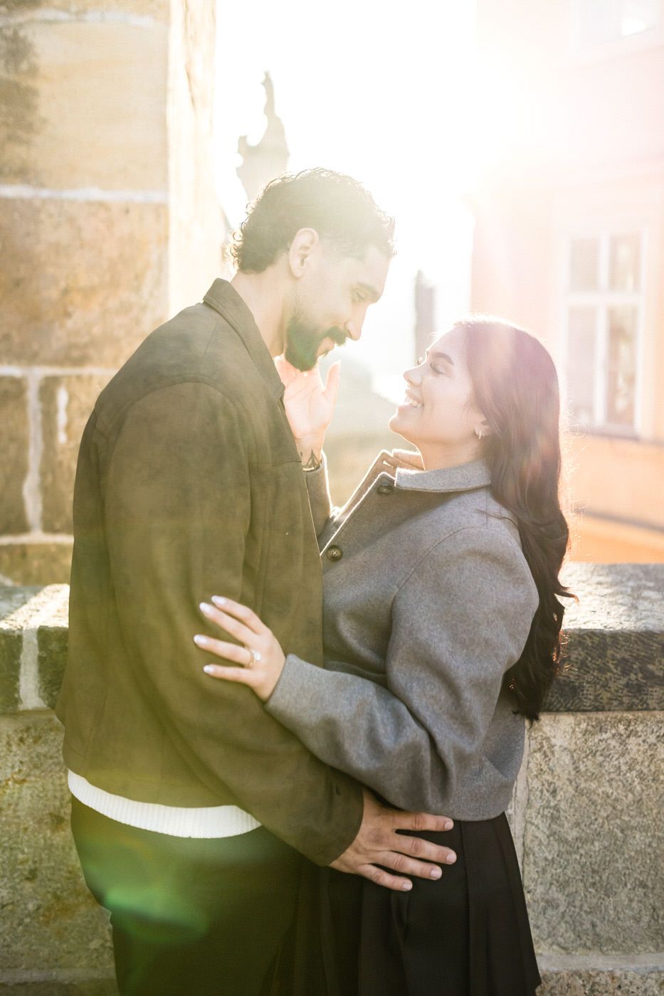 Smiling couple embracing near a stone wall in warm golden sunlight, the man in an olive jacket and the woman in a gray coat.