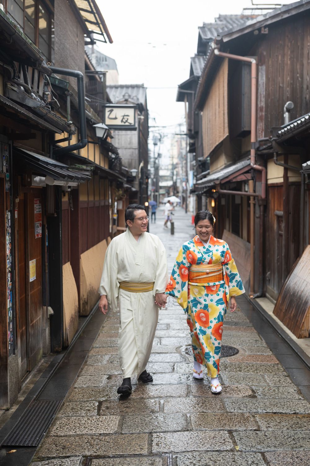 Couple in traditional Japanese kimono and yukata walking hand in hand along a stone-paved street lined with historic wooden buildings.