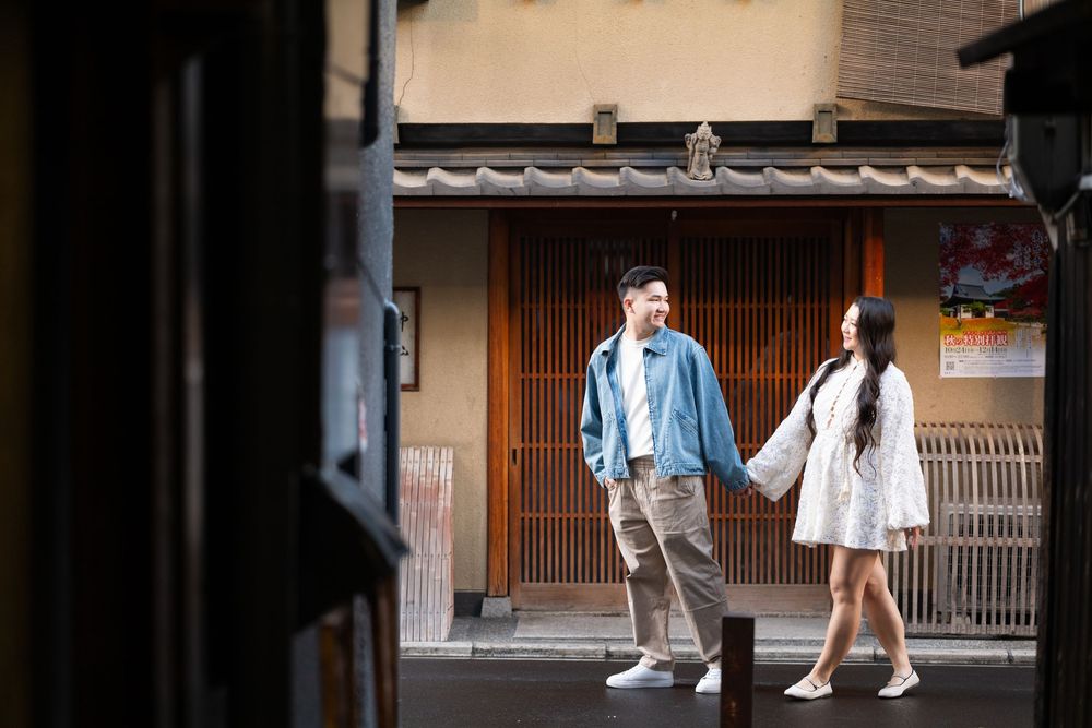 A couple holds hands while walking down a quiet traditional street in front of a wooden Japanese-style building.