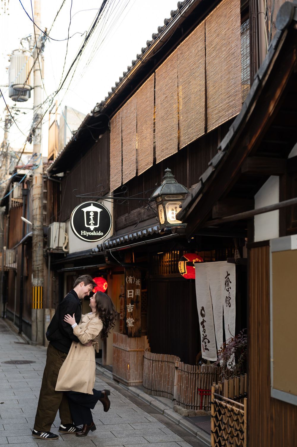 Couple embracing in a romantic pre-wedding photo on a traditional wooden street in Gion, Kyoto, with lanterns and noren curtains.