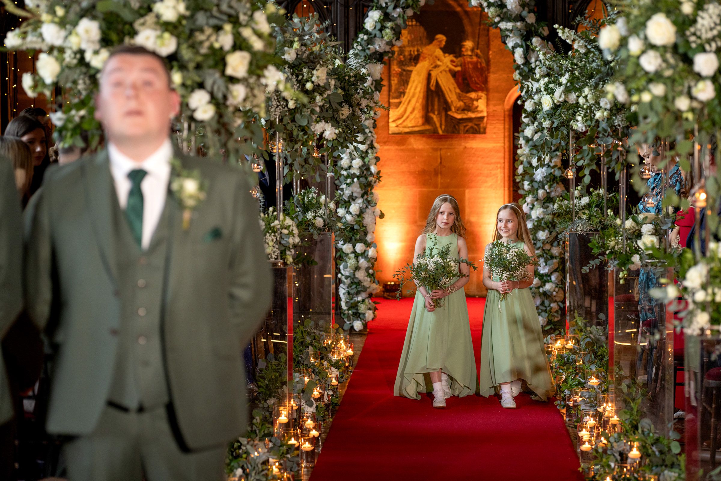 Groom in a green tweed suit stands as two flower girls in sage dresses walk down a red-carpeted aisle lined with white flowers in a church.