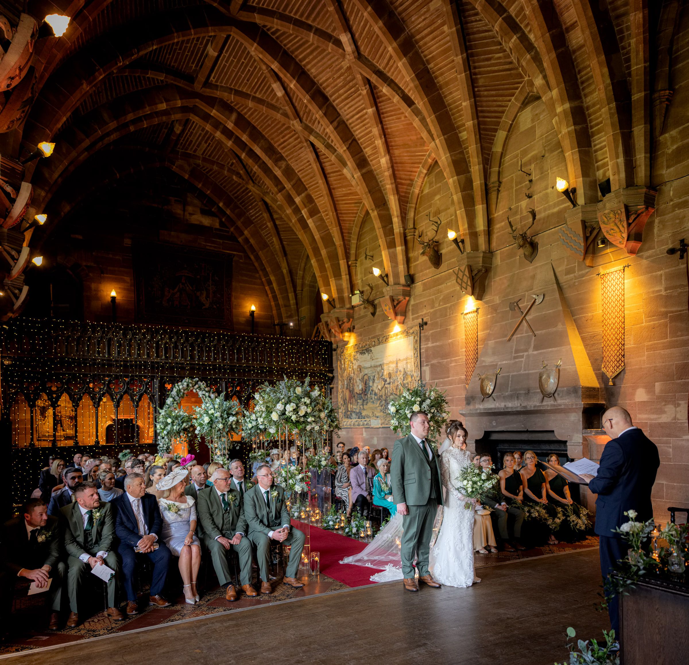 Bride and groom exchange wedding vows before seated guests in a historic stone hall with grand timber vaulted ceilings.