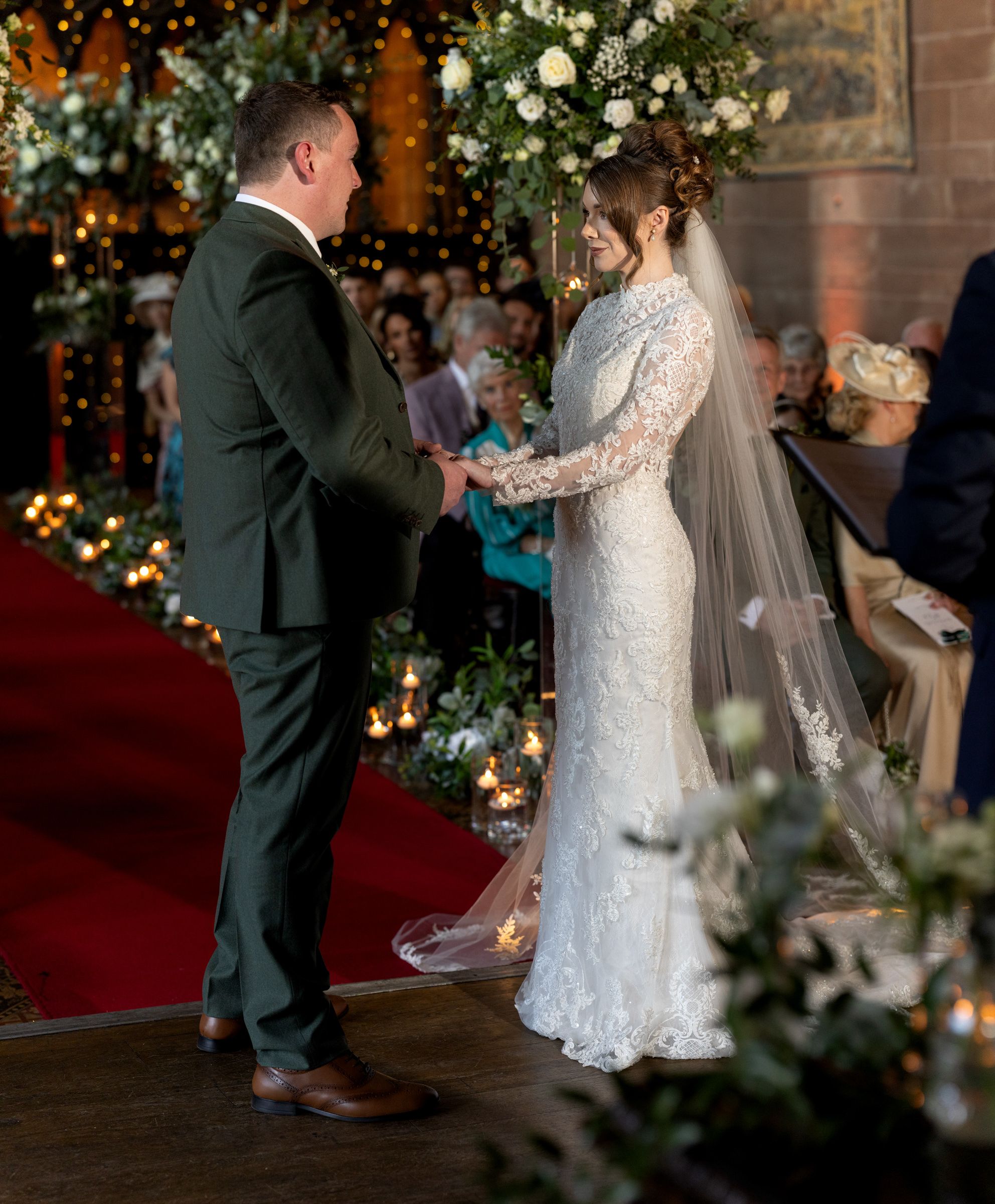 Bride in a lace gown and long veil holds hands with the groom during a candlelit wedding ceremony, with guests seated in the background.