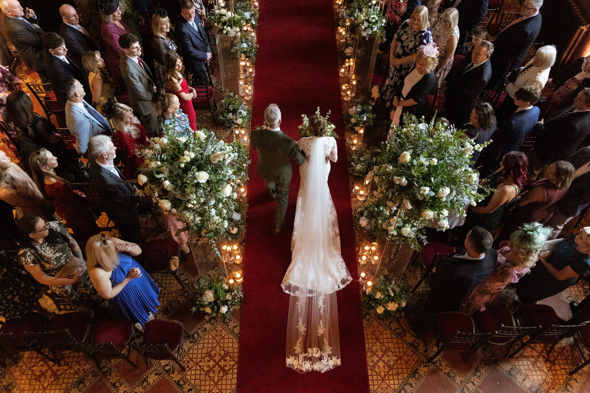 Overhead view of a bride in a long veil walking down a red-carpeted aisle between floral arrangements as wedding guests watch.