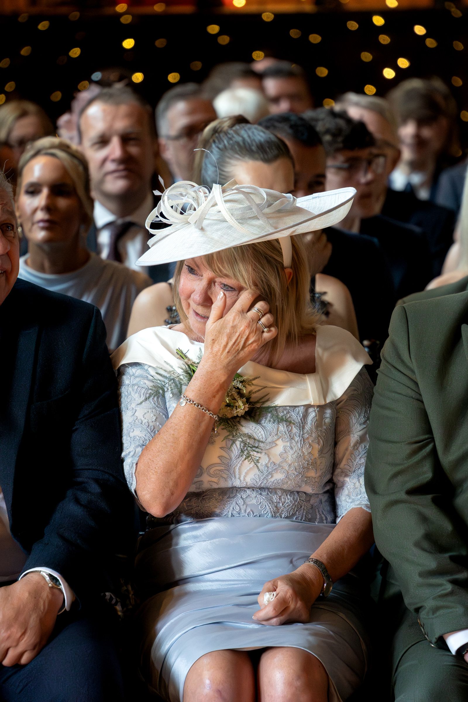 Emotional woman in a white fascinator and blue lace dress wipes a tear while seated among wedding guests under warm string lights.