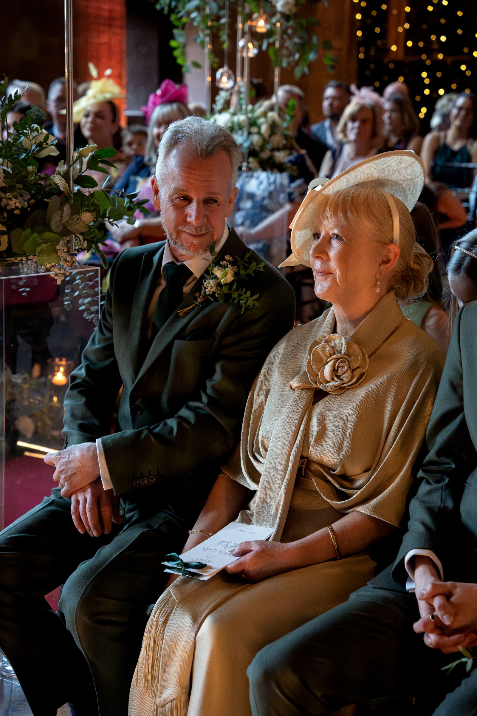 Middle-aged man in a dark suit with a floral boutonniere sits beside a woman in a champagne outfit and fascinator at a wedding ceremony.
