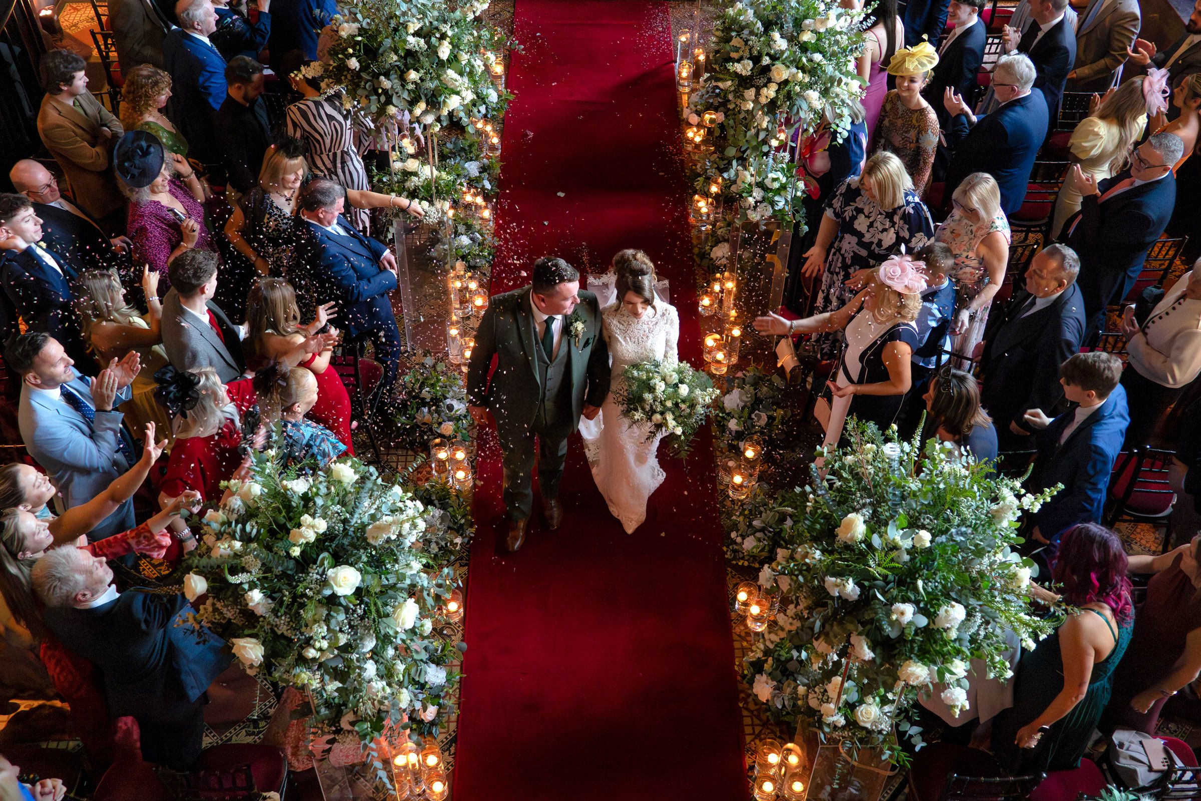 Bride and groom walk down a red carpet aisle lined with white floral arrangements as wedding guests watch from both sides.