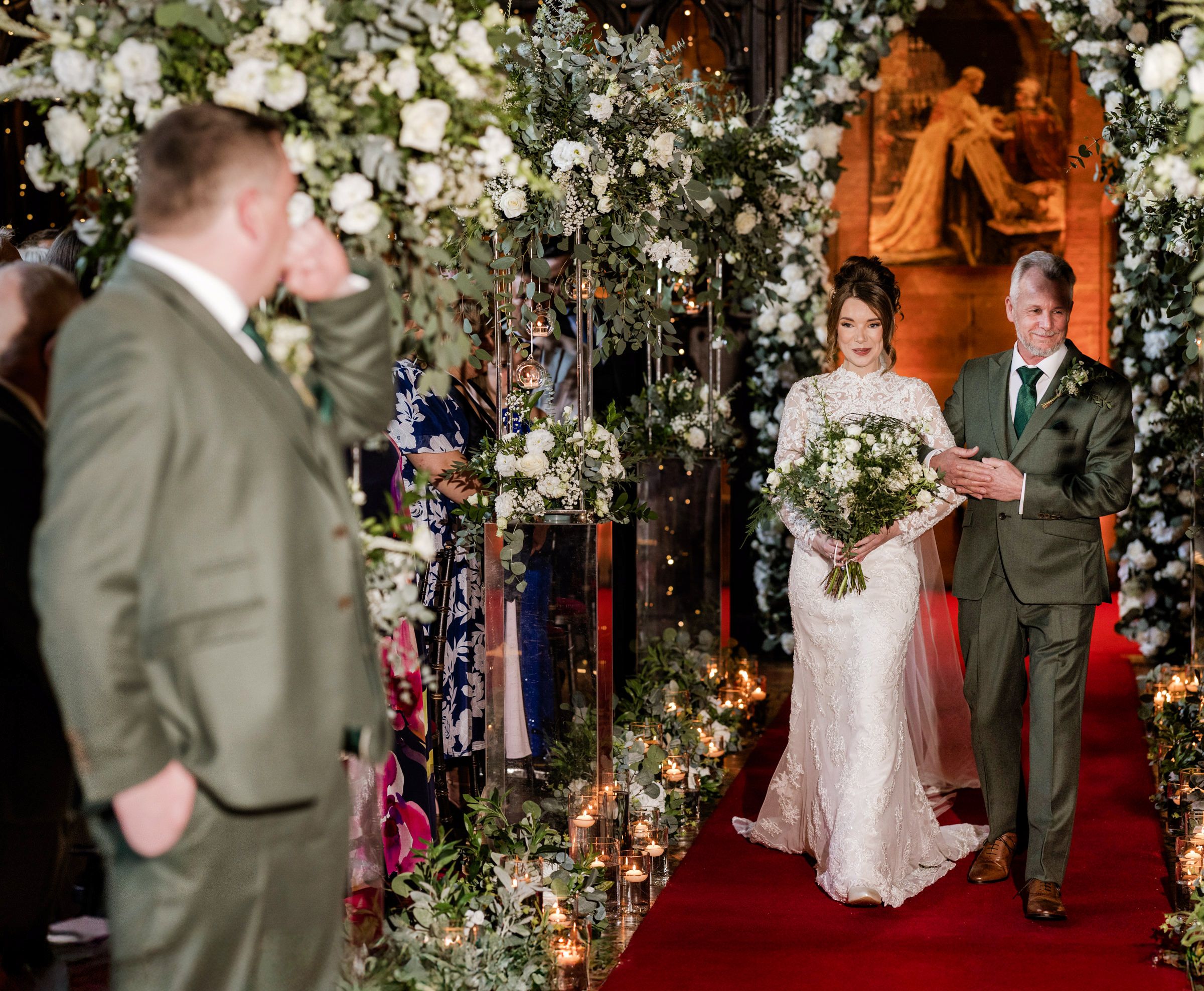 Bride in a lace gown carrying a white bouquet walks down a red-carpeted aisle with her father in a floral-decorated church.