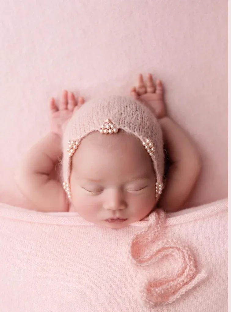 Sleeping newborn baby in a pink knit bonnet with pearl details, tucked under a soft pink blanket with tiny hands raised.