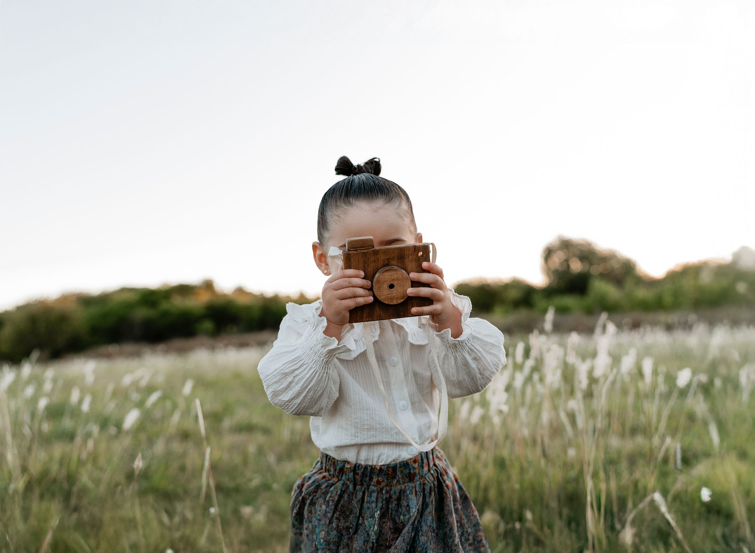 Young girl holding a vintage wooden toy camera in a green meadow field on a cloudy day.