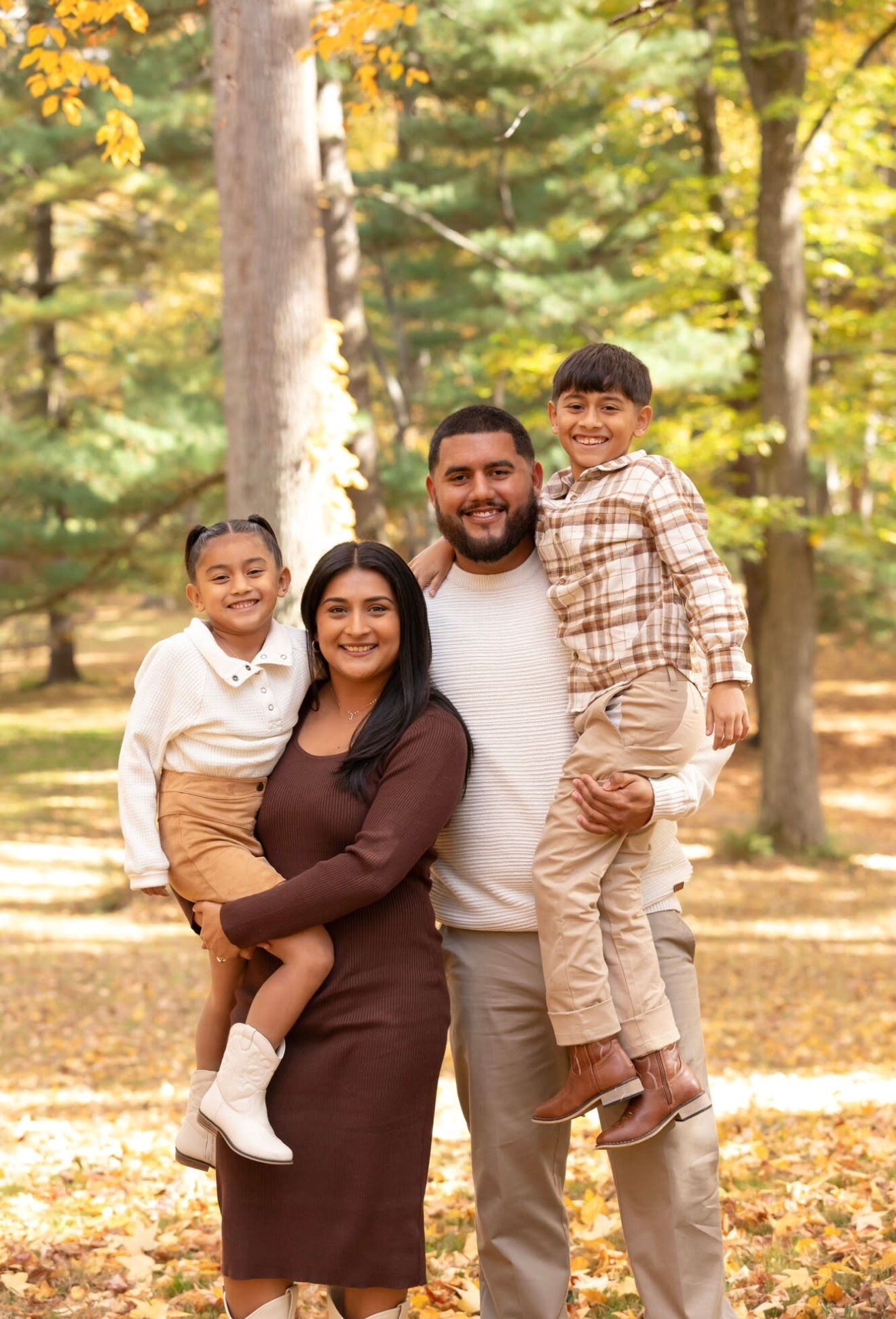 Family of four posing in autumn forest, parents holding two young children, wearing coordinated fall outfits in warm neutral tones.