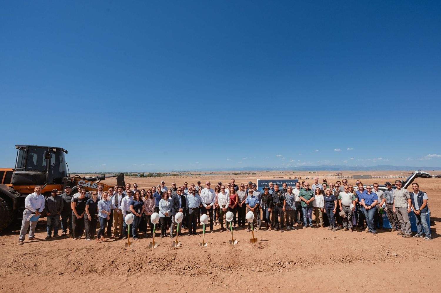 Large group of people gathered on a construction site under bright blue sky during groundbreaking ceremony.
