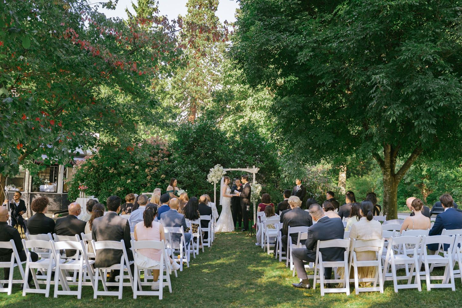 Outdoor garden wedding ceremony with guests seated in white chairs on a lush green lawn beneath canopy trees.