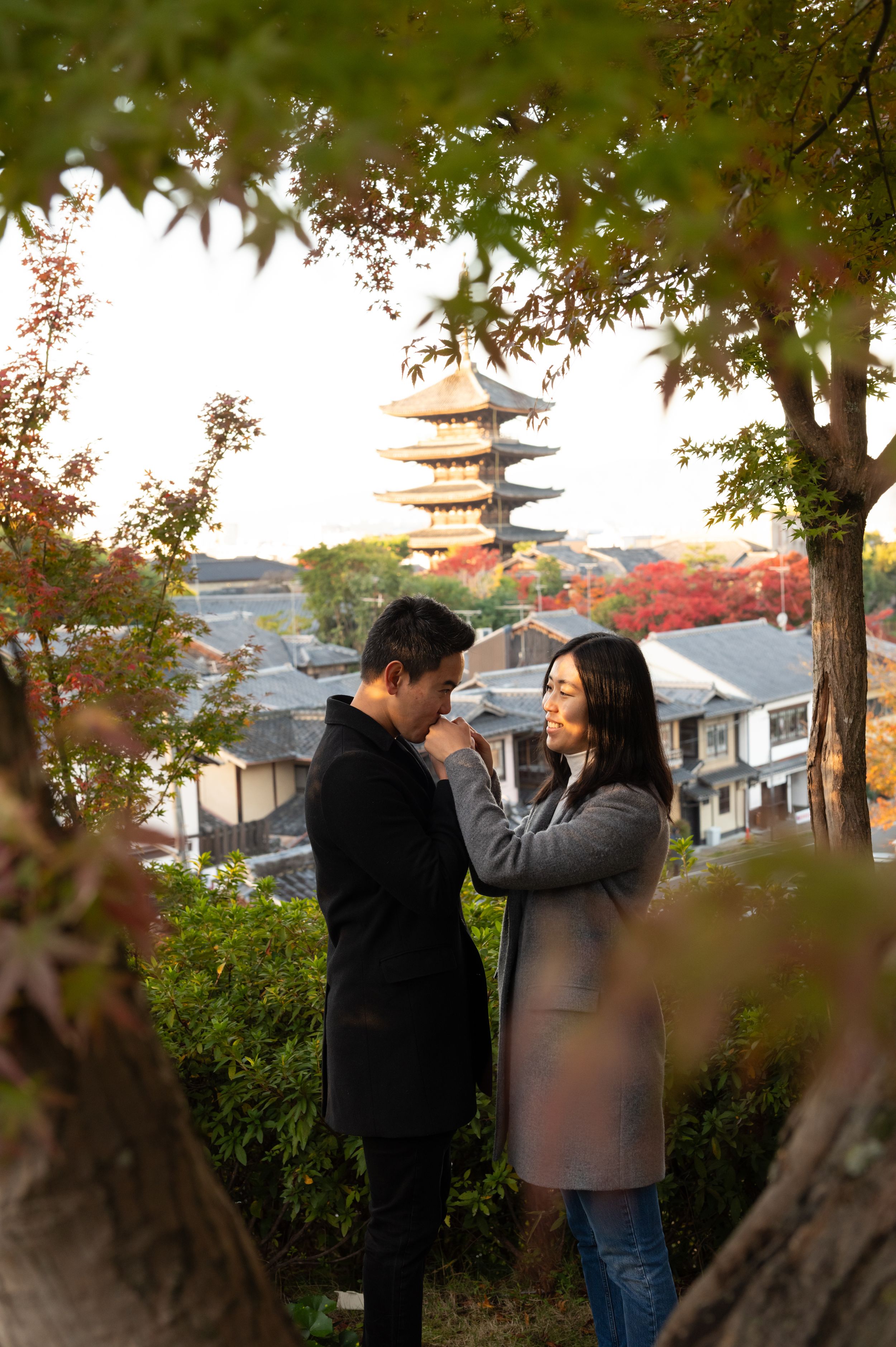 Couple shares an intimate moment among autumn foliage, overlooking Kyoto rooftops and a historic multi-tiered pagoda at sunset.
