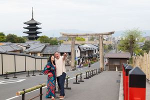 Couple in traditional kimono takes a selfie on a Kyoto street with a stone torii gate and five-story pagoda in the background.