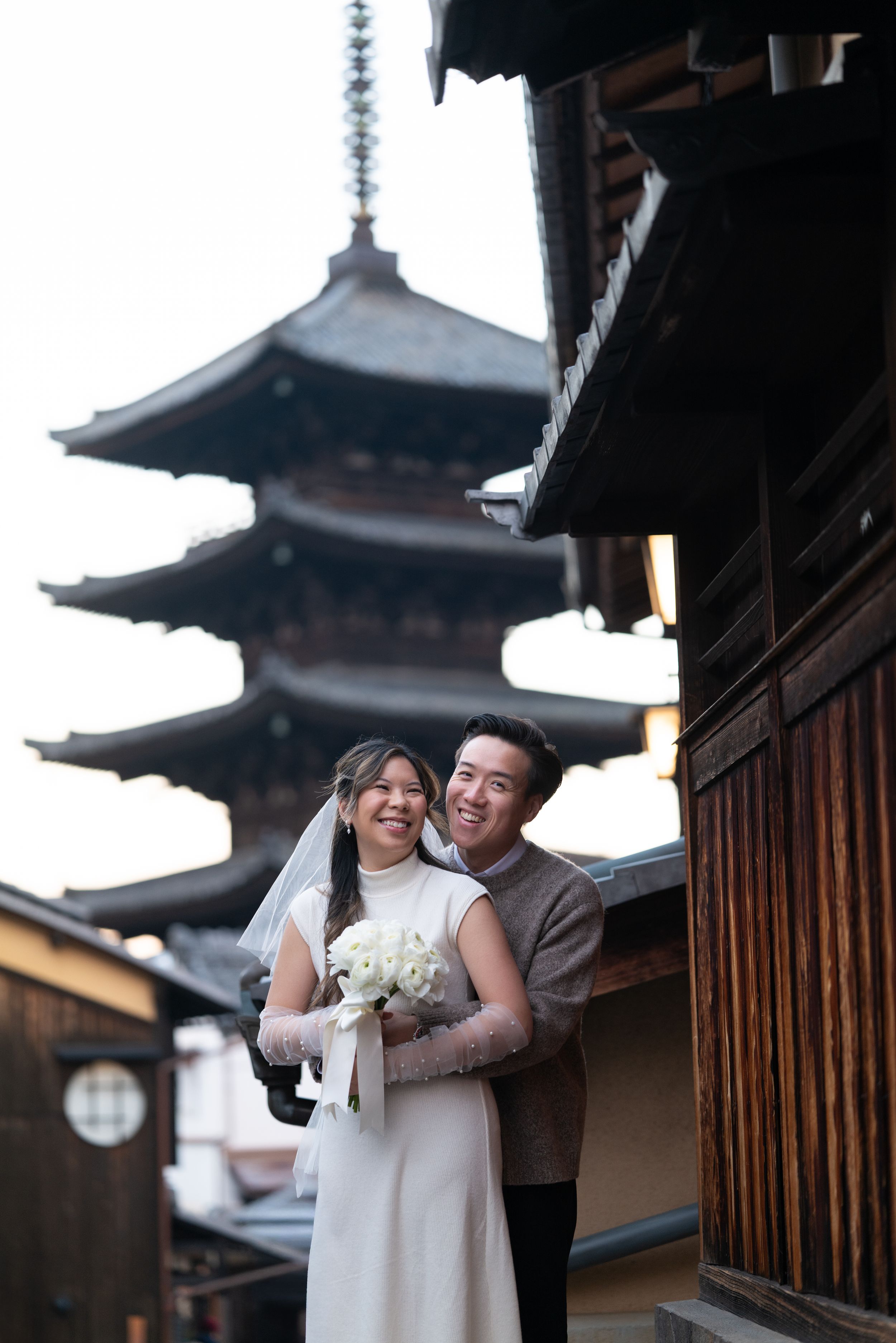Smiling bride in a white dress holding a bouquet is embraced by her groom beside traditional wooden buildings and a Japanese pagoda.