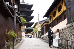 Bride in white gown and groom in dark suit pose on a quiet stone-paved street in Kyoto with a traditional wooden pagoda behind.