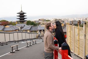 Couple kissing on a red railing overlooking a Kyoto cityscape with a traditional five-story pagoda and distant mountains at dusk.