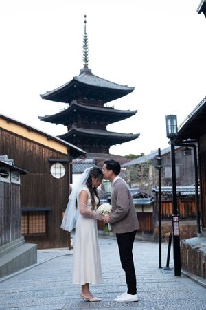 Bride in a white dress and veil holding a bouquet stands close to a groom on a quiet Kyoto street with a pagoda behind them.