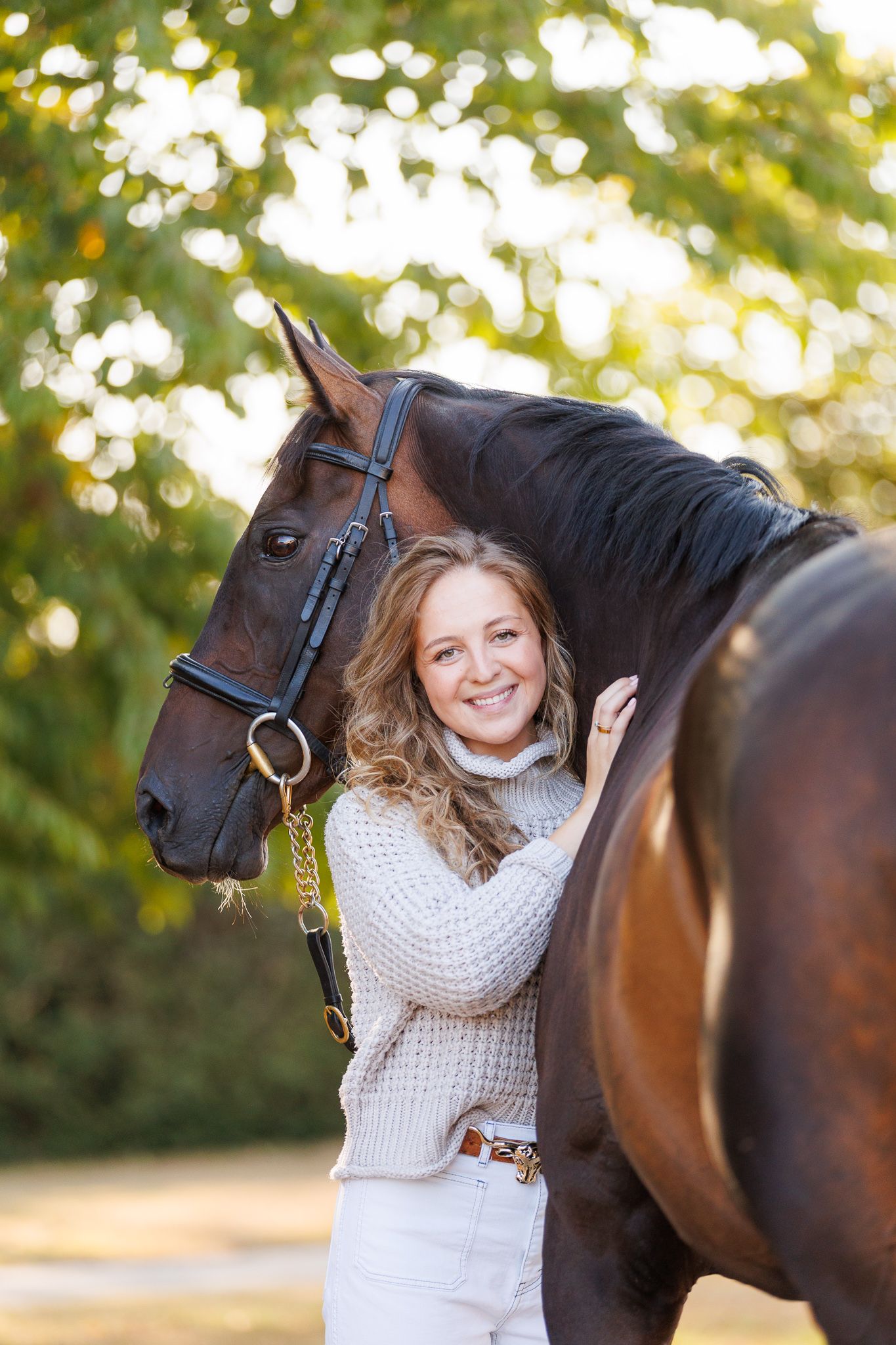 Young woman smiling between two bay horses with white markings, holding reins near a brick stable.