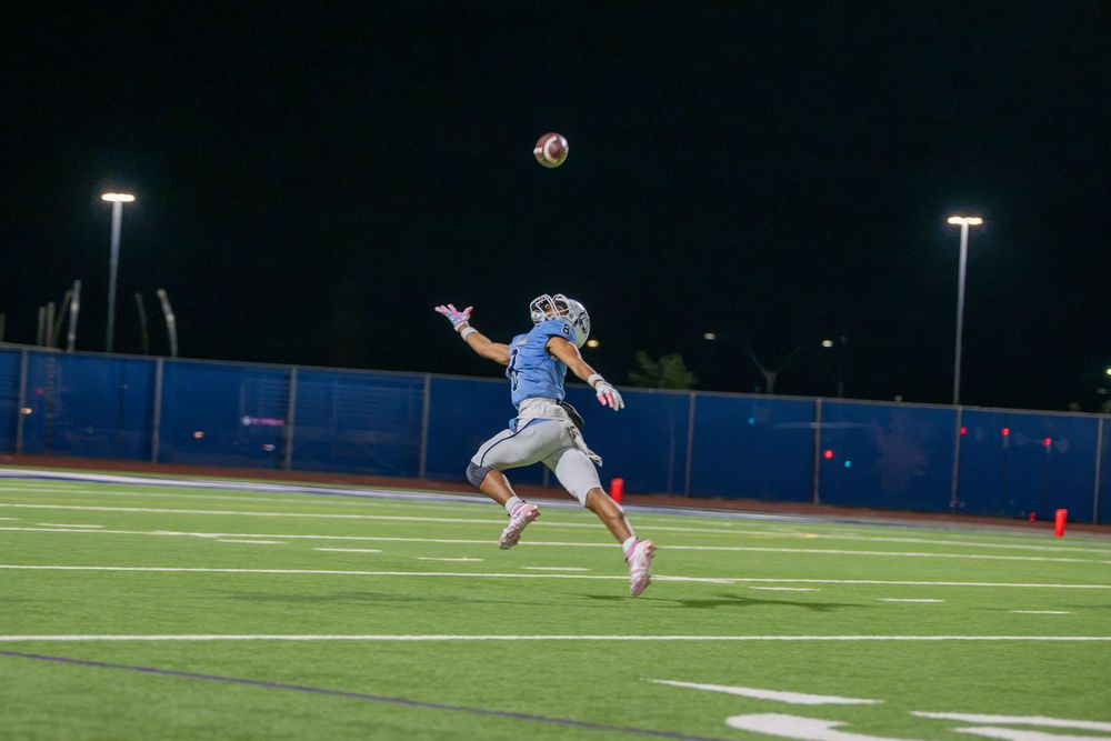 A high school football receiver in a light blue jersey leaps to catch a pass during a night game on a lit stadium field.