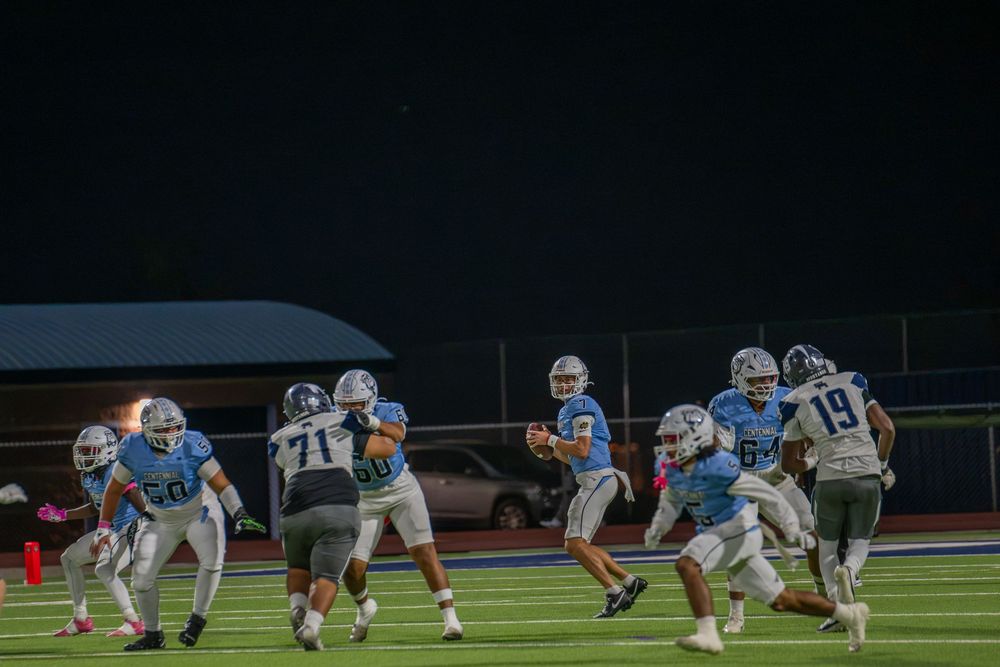 High school football players in light blue and white uniforms run a play under stadium lights as the quarterback looks to throw.
