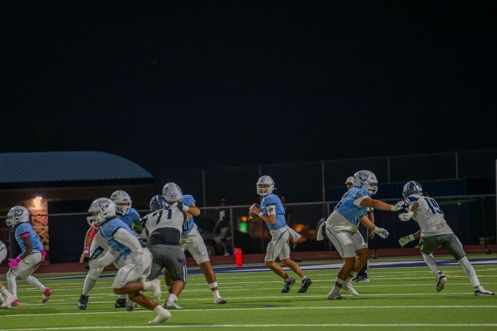 A quarterback in a light blue jersey drops back to pass as teammates and defenders in white clash on a lit football field at night.