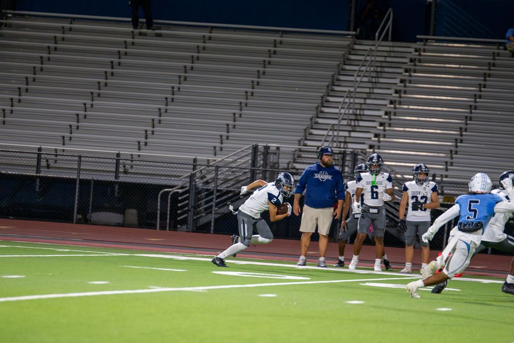 High school football players in white and blue uniforms run drills on a green field while a coach watches near empty stadium bleachers.
