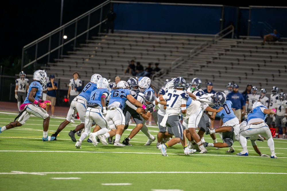 Football players in blue and white uniforms collide in a crowded tackle during a night high school game as fans watch from the stands.