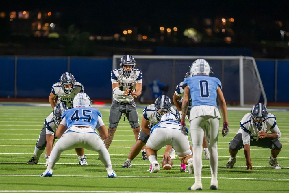 High school football players in white and blue uniforms line up at the line of scrimmage during a night game on a lit stadium field.