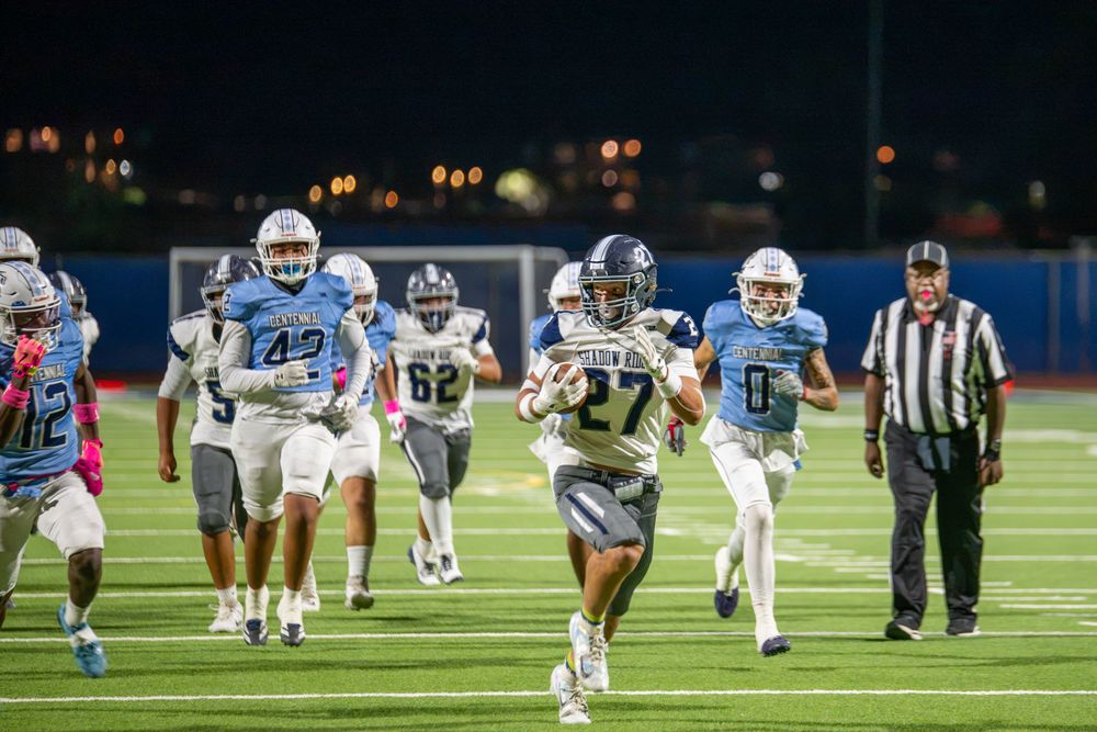 A running back in a gray and navy uniform sprints downfield with the football while light blue defenders chase and a referee watches.