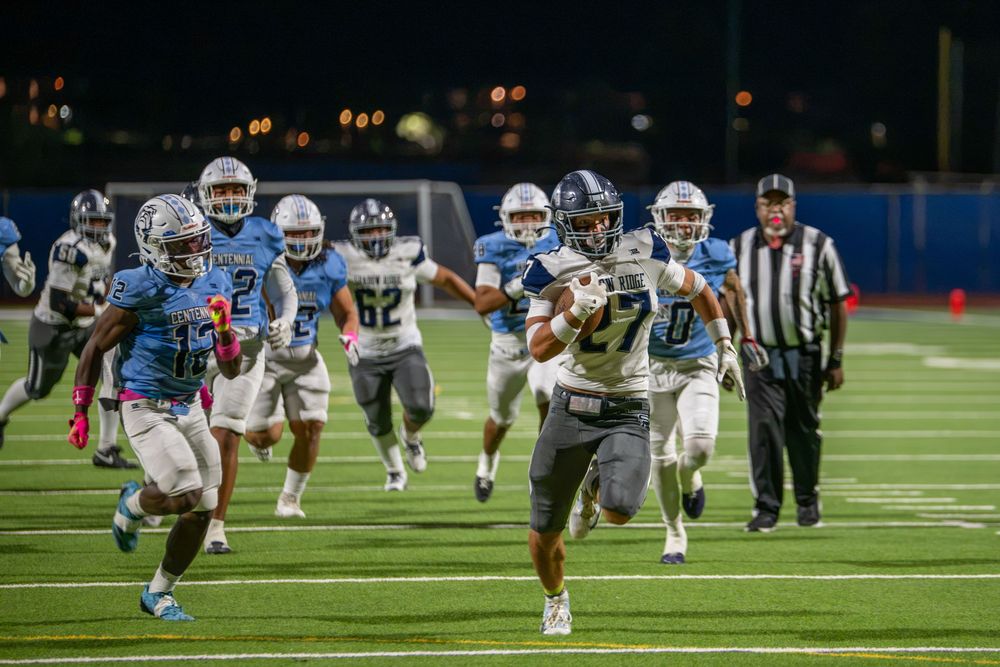 A football player in a gray and black uniform sprints down the field with the ball as defenders in blue chase him during a night game.