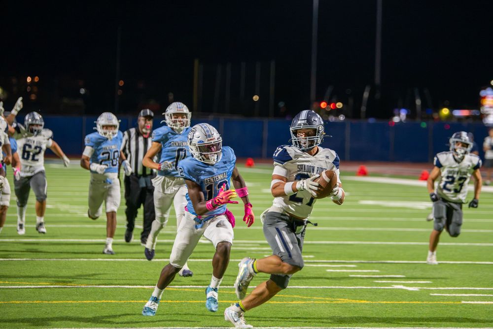 A ball carrier in a white and navy uniform runs with the football past a diving defender during a Friday night high school football game.