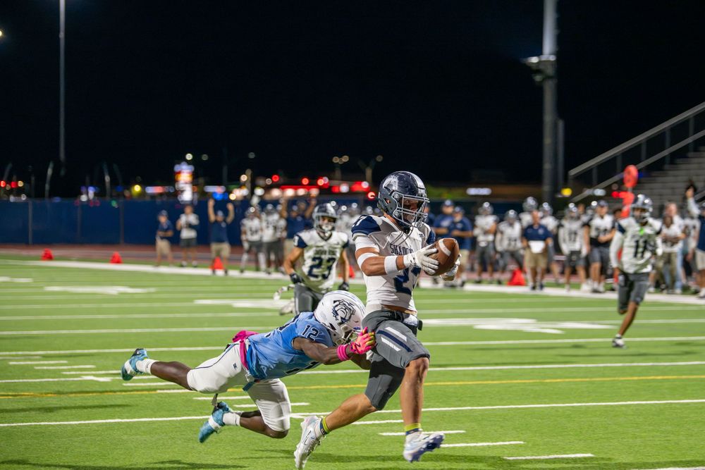 A football player in a gray uniform carries the ball downfield while a defender in light blue dives to tackle him during a night game.