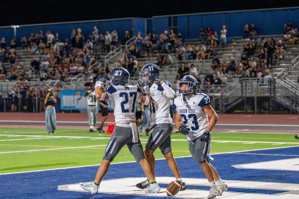 Three football players in white and navy uniforms celebrate on the field during a night game as fans watch from the stadium bleachers.
