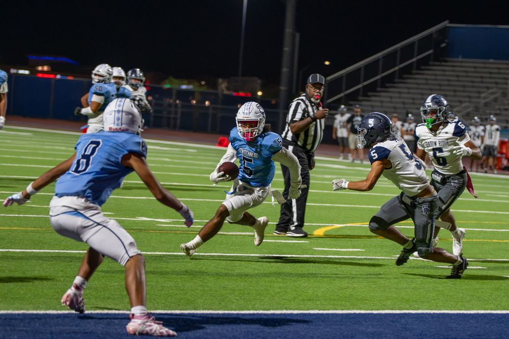 High school football player in light blue uniform carries the ball toward the end zone as defenders in white chase during a night game.