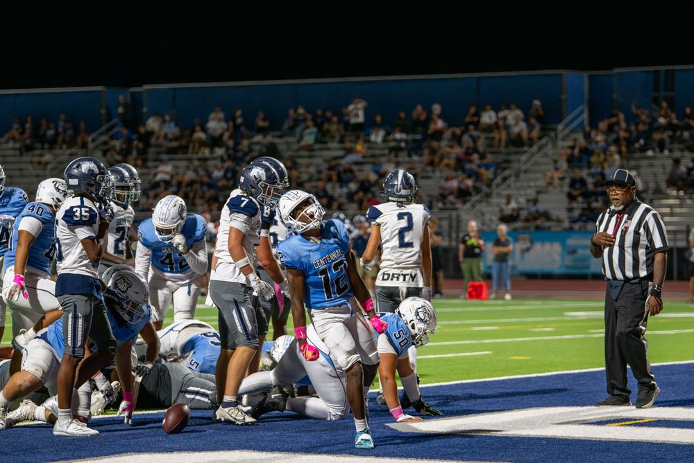 Football players in light blue and white uniforms pile up near the end zone as a referee watches during a high school night game.